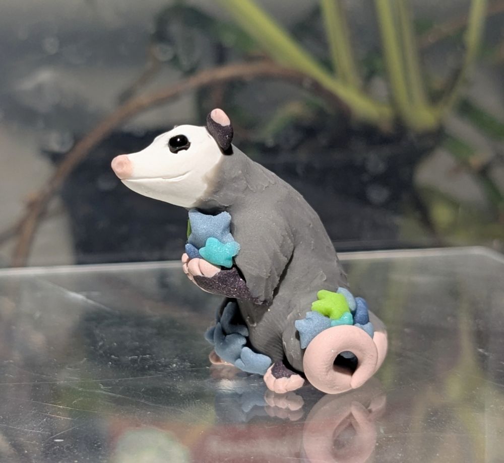 A figurine of a stylized opossum holding a collection of five pointed stars in his arms, and tail. He sits back on his haunches, and looks happy. 