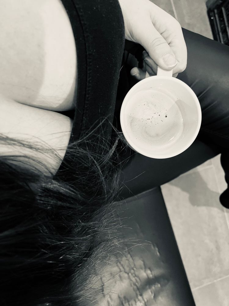 Schwarz-weiß Bild einer Frau die von oben fotografiert wird. Im Fokus liegen ihre Brüste in einem tief ausgeschnittenen Top sowie eine helle Tasse mit Kaffee. Sie sitzt auf einer schwarzen Bank.