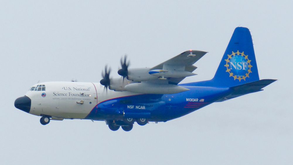A white/blue C-130 with the logo of the National Science Foundation prominently featured on the vertical stabilizer makes a low approach in the landing configuration 