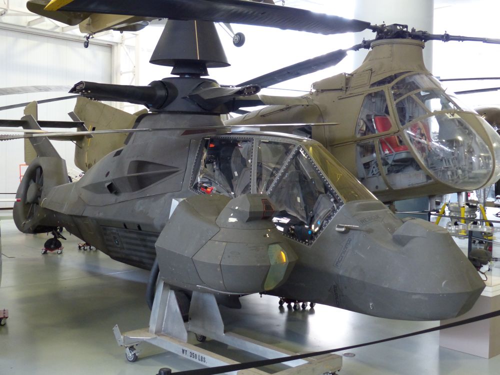 RAH-66 Comanche, a stealth attack helicopter, viewed from the front and to the side on display at a museum. A nose radome is sitting on a stand beside it. The main rotor blades have been removed.