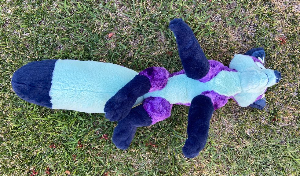 Purple and mint green fox plushie lying on its back on grass