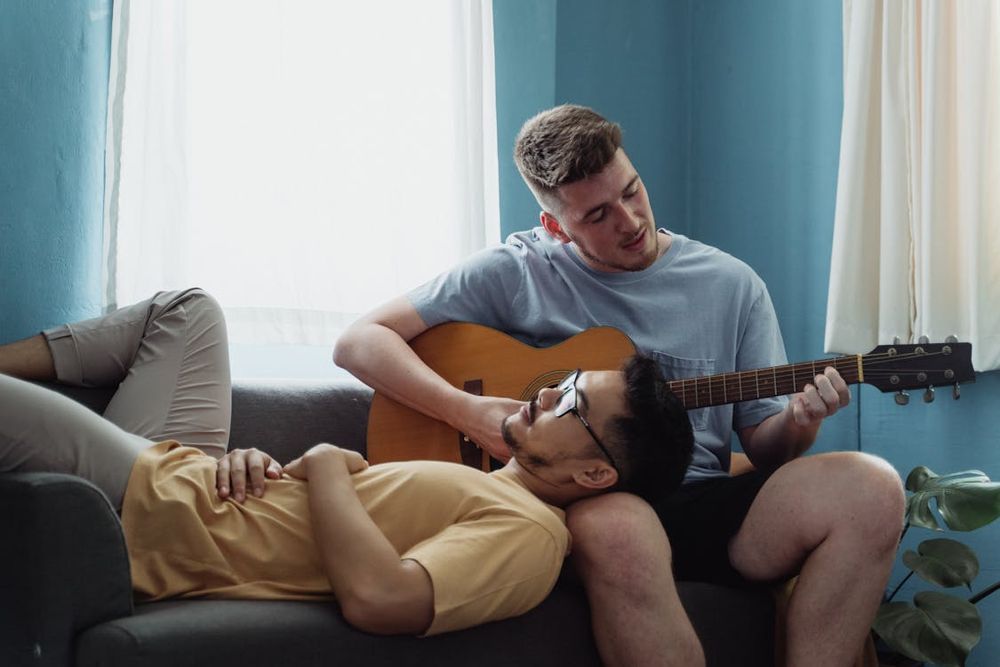 The guitar is now in the hands of the second man. He strums calmly, focused on his work. The first man lays on the sofa beside him and rests his head in the other's lap, arms laid across his torso. It is intimate and soft in the bright and airy room.