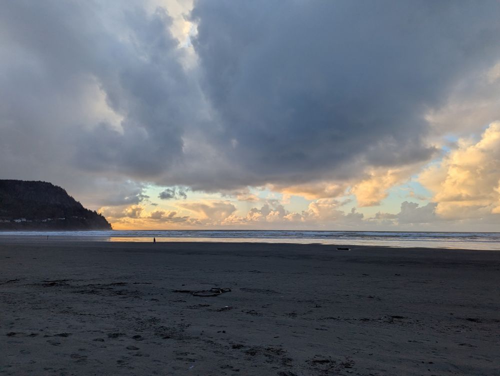 another Oregon coastal beach, at sunset. there is an impossibly large stretch of flat sand with a sliver of the ocean across the horizon. a distant bluff on the left edge is covered in pine trees, and has a house-lined street gently curving up it. the sky is awash in pastel blues and orange, with angry grey storm clouds rolling in.