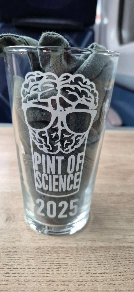 A pint glass with the Pint of Science logo (a brain wearing a pair of glasses, itself sitting in a pint glass), and 2025 underneath. A grey tote bag has been shoved inside the glass to make the logo stand out better.