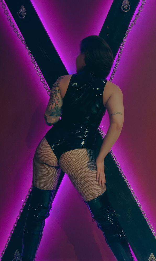 A tattooed woman in black PVC body and thigh high boots, standing in front of a BDSM cross. The cross is black and set against a red wall. There is pink light shining under the cross.