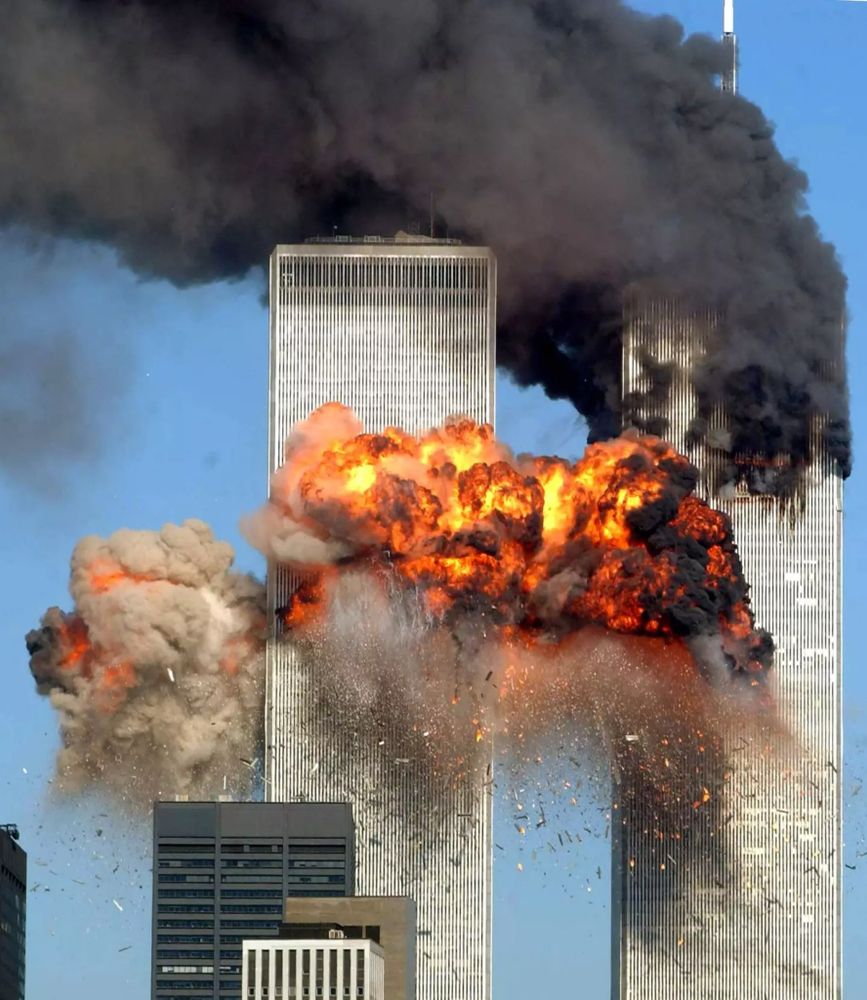 Twin towers shortly after impact on 9/11/2001. Black smoke is billowing from the top of the right hand tower and blowing to the left. Orange and yellow fireballs with black edges are coming out of the left hand tower part way below the top.
