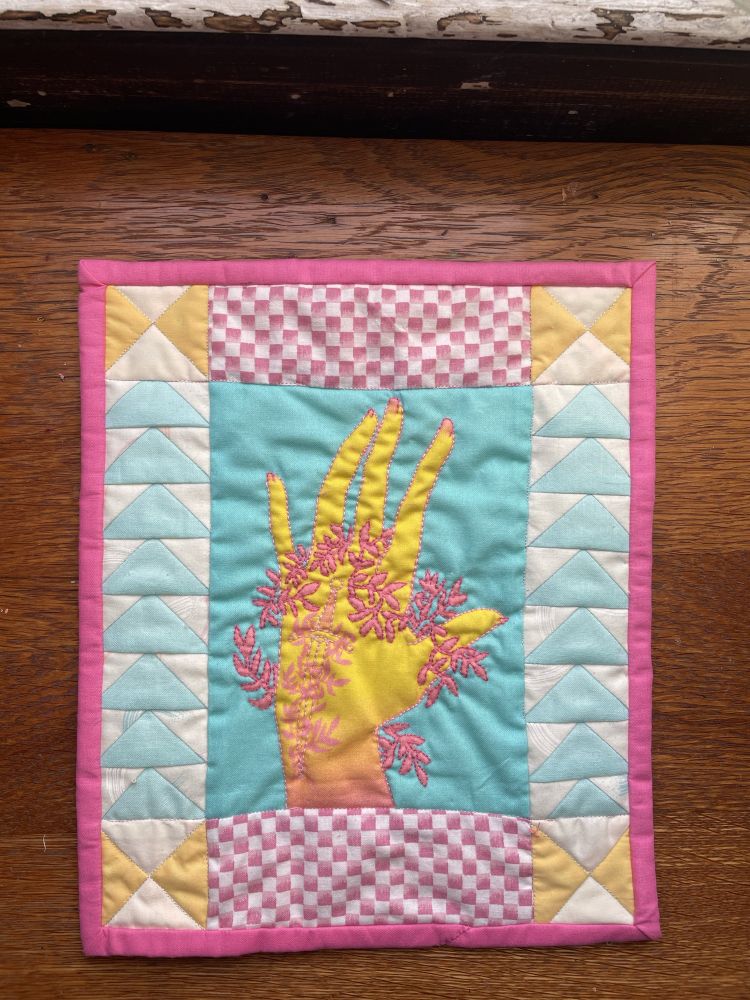 Photo of a small quilt wall hanging. The center of the quilt is printed with a drawing of a yellow hand surrounded by pink vines; the hand has a tattoo of a sword. On each side of the centerpiece is a row of flying geese pointing upward. Above and below the centerpiece are strips of pink and white checkered fabric. The corner pieces of this quilt are quarter square triangle blocks.