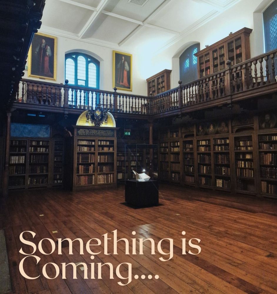 Photograph of Cosin’s Library with the text “Something is Coming” in the bottom left corner. Library has wooden floors with bookcases along the walls, a balcony, two portraits and large windows. A museum case with a book on display is in the centre of the library.
