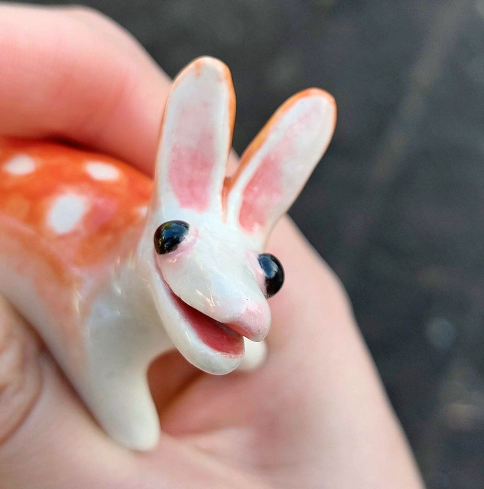 a small ceramic animal held in my hand. it has a gentle yet exuberant smile, round shiny black eyes, a snout with no nose or teeth and tall rabbit-like ears. it has pink accents on its white face and a soft-edged light red upperside to its body, with white spots. its feet and underside are white.