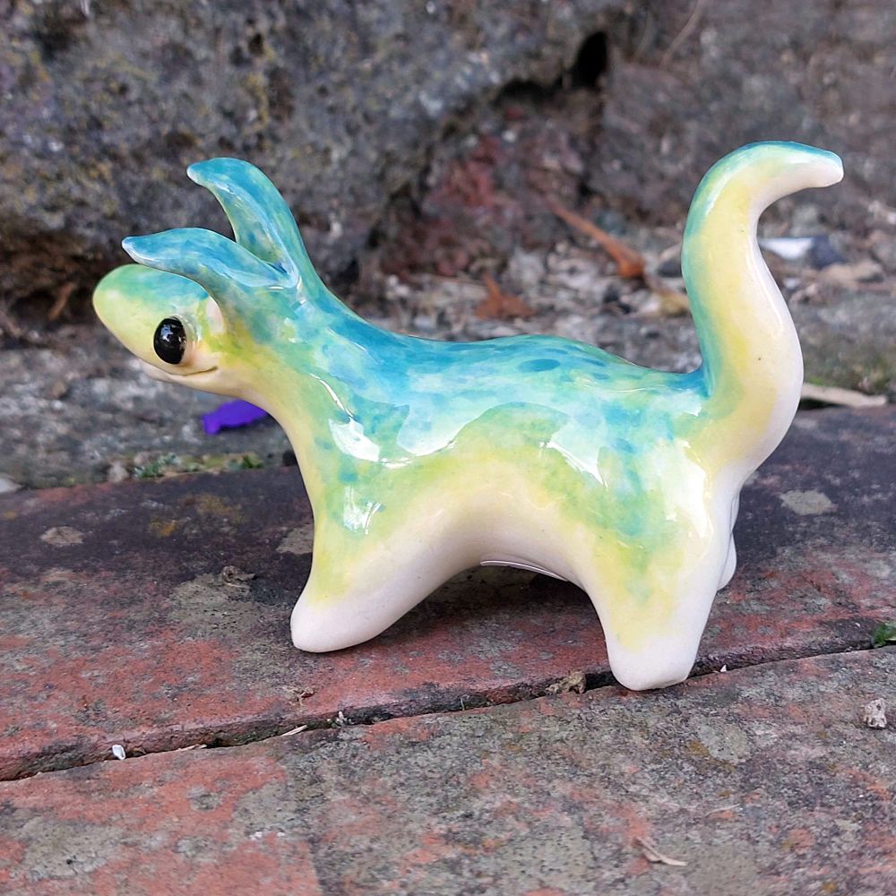 The same ceramic creature shown from one side. A teal-green-yellow-white gradient can be seen along its body, with its underside being white and its upper side being teal. Its tail has a feline S-shape.