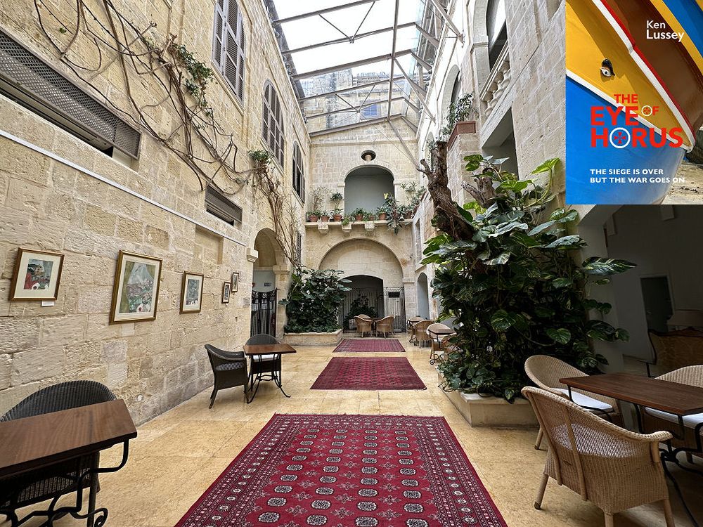 This modern image shows the interior of the Xara Palace Hotel in Mdina, Malta. A broad corridor with sumptuous red rugs extends out to the middle of the frame. It has light stone walls that extend upwards over two floors to a sloping glass roof. There are tables and rattan chairs at intervals along the sides of the corridor and large displays of green plants. The front cover of ‘The Eye of Horus’ is shown in the top right corner.