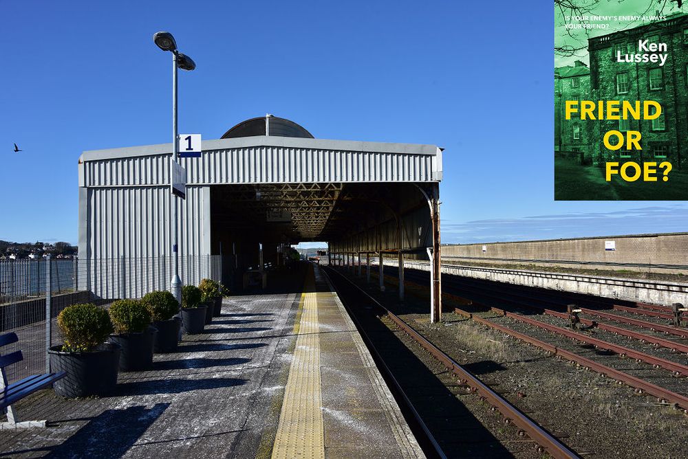 This modern image shows Stranraer railway station. A platform extends into the frame from the bottom left corner with a chain link fence and potted plants to its left. To its right are four railway lines. The platform and left-hand line are enclosed by a crinkly shed which occupies the middle left of the frame. The sea can be glimpsed on the left and the sky is blue. The front cover of ‘Friend or Foe?’ is shown in the top right corner.