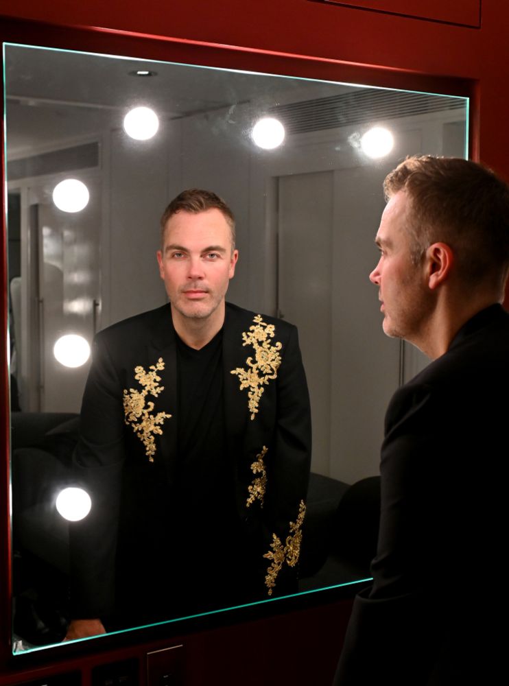 Nicholas McCarthy wearing a black jacket with gold decoration, looking in a Royal Albert Hall dressing room mirror lit by bulbs