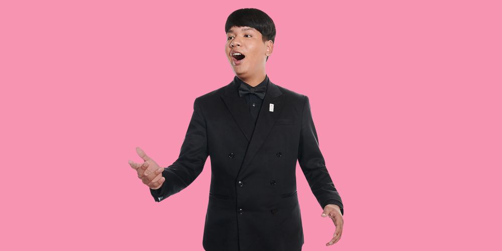 A man wearing a black suit singing in front of a pink background