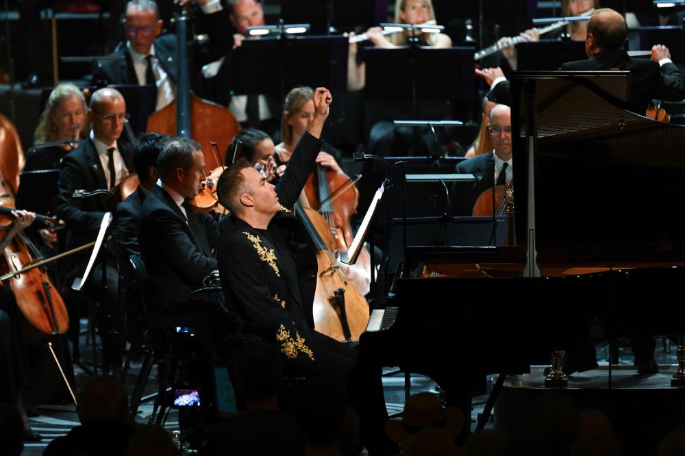Nicholas McCarthy at a grand piano in the Royal Albert Hall, arm in the air, orchestral musicians behind him