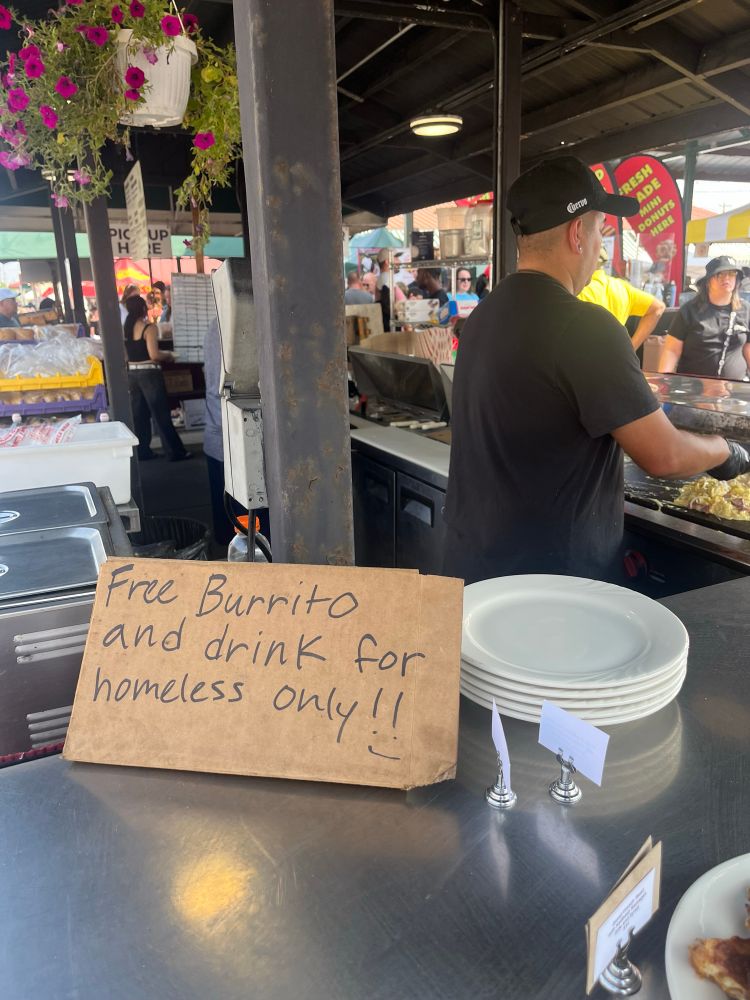 A Mexican food station at the farmer’s market with a cardboard sign that says “free burrito and drink for homeless only”