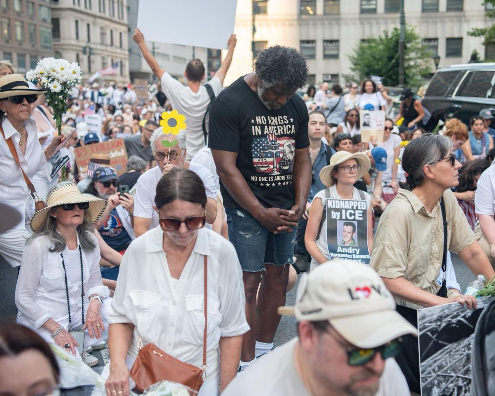 Protesters kneel and sit outside of 26 Federal Plaza. One person in a “No Kings in America” T-shirt stands, staring at the ground in a moment of silence.