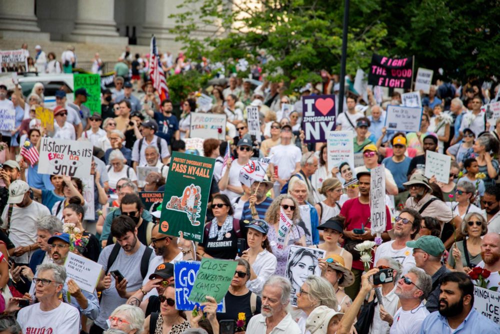 Tens of thousands of protesters gather in Foley Square, NYC for Good Trouble Lives On protest. There are many signs and American flags.