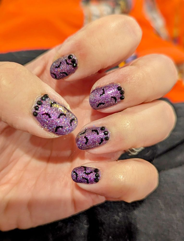 A close-up of my new manicure. The nails are painted with Holo Taco's "Crystal Mommy," a purple holographic polish. Little bat confetti is scattered over each nail, and a string of black gems rests at the base of each nail.