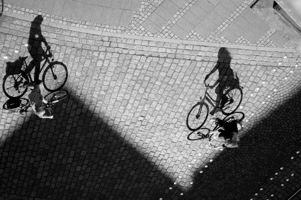 Zwei Radfahrer:innen fahren auf einer Straße, die mit Kopfsteinpflaster bedeckt ist. Die Szene ist aus der Vogelperspektive aufgenommen. Licht und Schatten formen pyramidenartige Flächen auf dem Boden. Die langen Schatten der Radfahrer sind deutlich zu sehen. Durch die Schwarz-Weiß-Fotografie wird das Spiel mit Licht und Schatten betont.