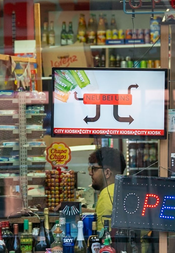 Innenansicht eines Kiosks mit einem digitalen Bildschirm, der Werbung für neue Produkte. im Hintergrund ist ein Verkäufer in gelbem T-Shirt zu sehen.
