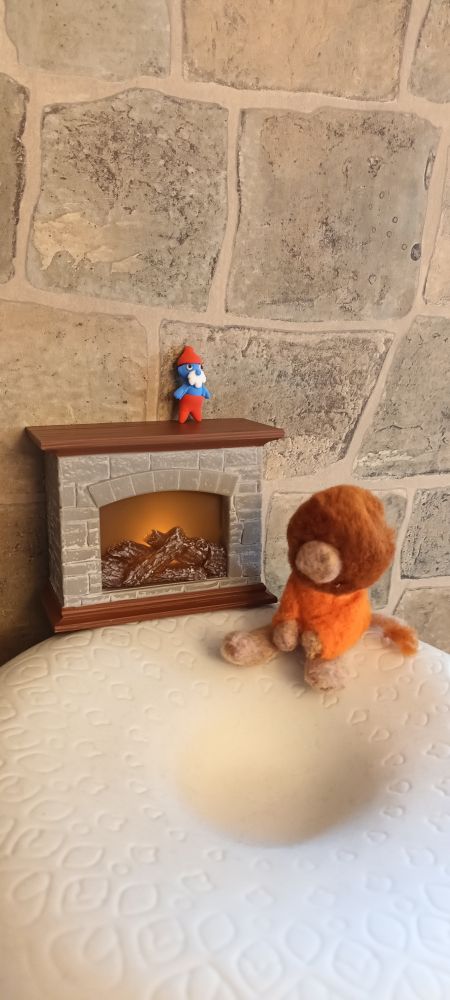 A nisse/Tontu wearing an orange sweater (all hand made from wool which has been needle felted by my youngest child) is sitting, warming himself beside an animated, battery operated mini yule fire with a tiny Papa Smurf doll on top of the hearth.
