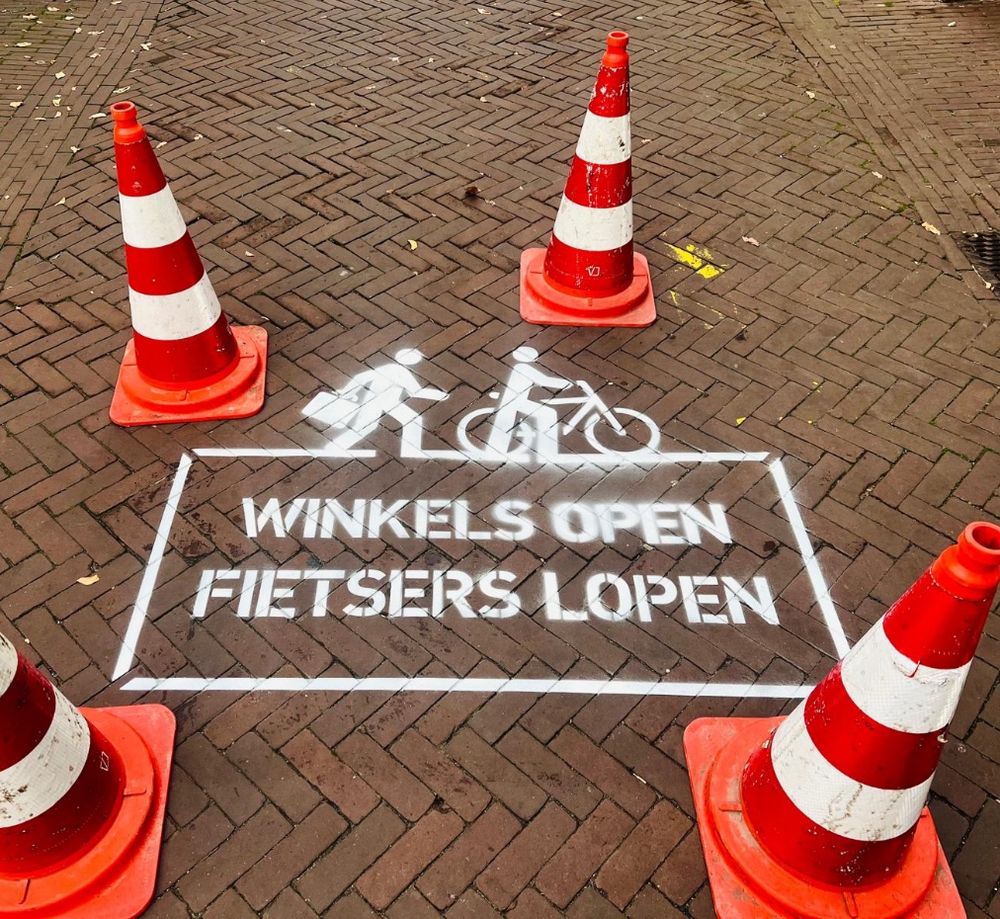 4 pionnen om een witkalk tekst op de stenen op de grond. Winkels open fietsers lopen. 