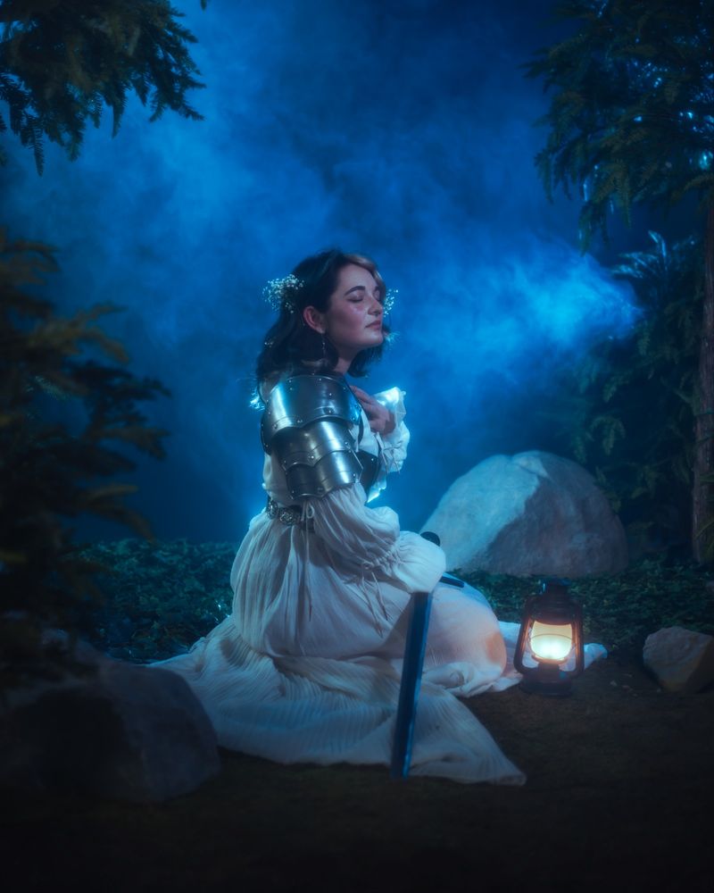 Girl I'm a white dress with armor sitting down with her eyes closed holding a sword. There's also a lantern