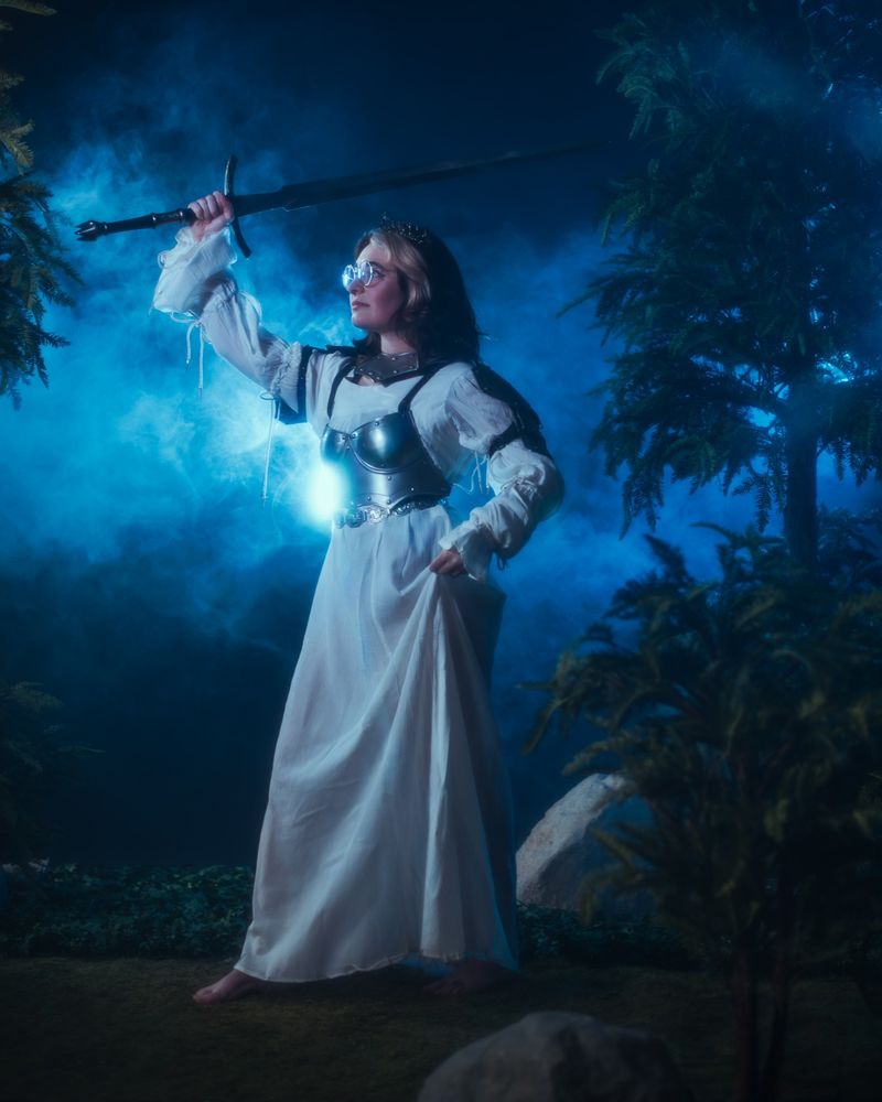 Girl in a white dress with armor holding up a long black sword.