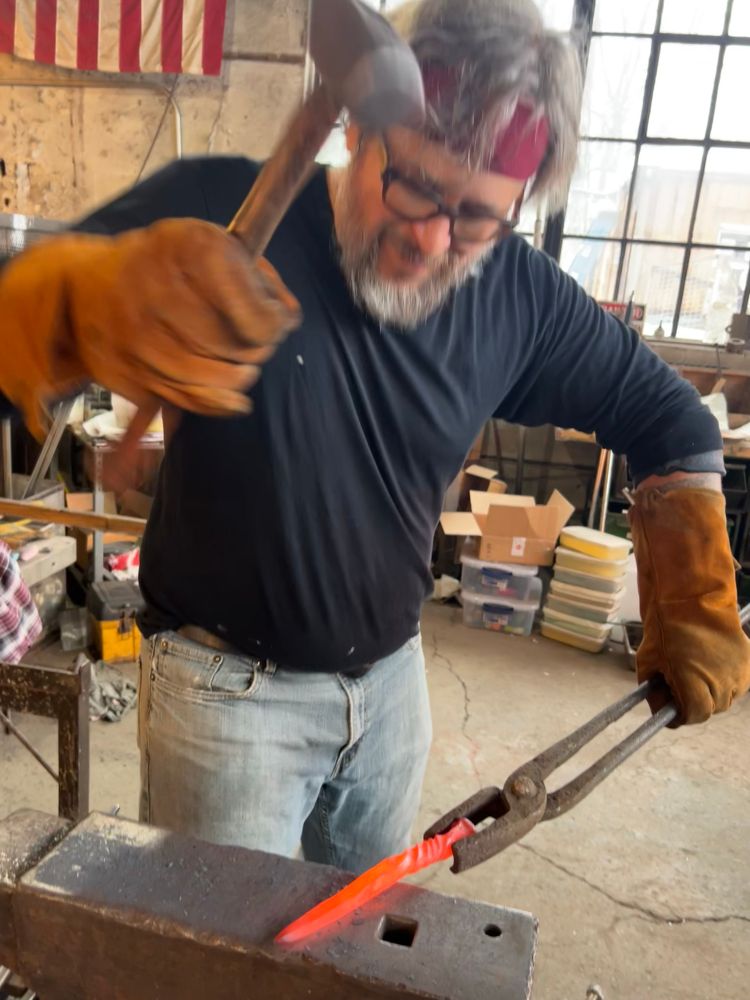 An old man, me, wearing a bandana and wielding a hammer on a hot orange piece of steel on an anvil. 