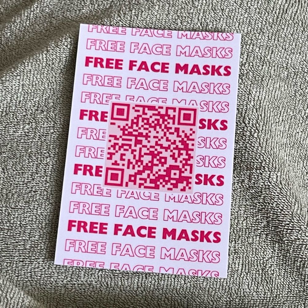 A photo of a rectangular sticker with a white background, that has pink text which reads “FREE FACE MASKS” repeatedly, with a dark pink QR code on top of the text. The sticker is resting on top of a green towel-like material. 