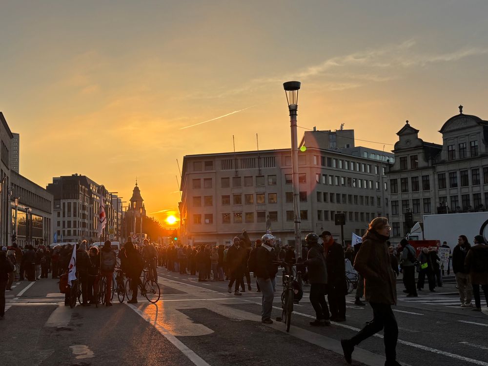 Sun setting in the distance over Brussels buildings with large crowd of strikers at a fairly central rally in silhouette in the foreground 