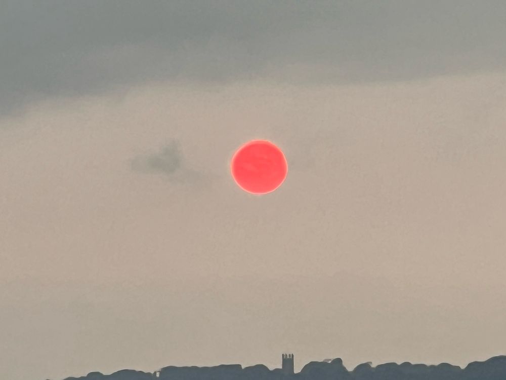 Greyish sky with cloud a darker grey across the top ~1/5th of the pic, a silhouette of the top of a cliff line/hills with the shape of a church tower discernible right at the bottom, and just above the centre midline the sun is basically a big red disc with a slightly yellow outline which looks like I cut it out of red paper and stuck it on the drew round it with a thin yellow marker pen. No “bleed” of colour like a usual sunset.