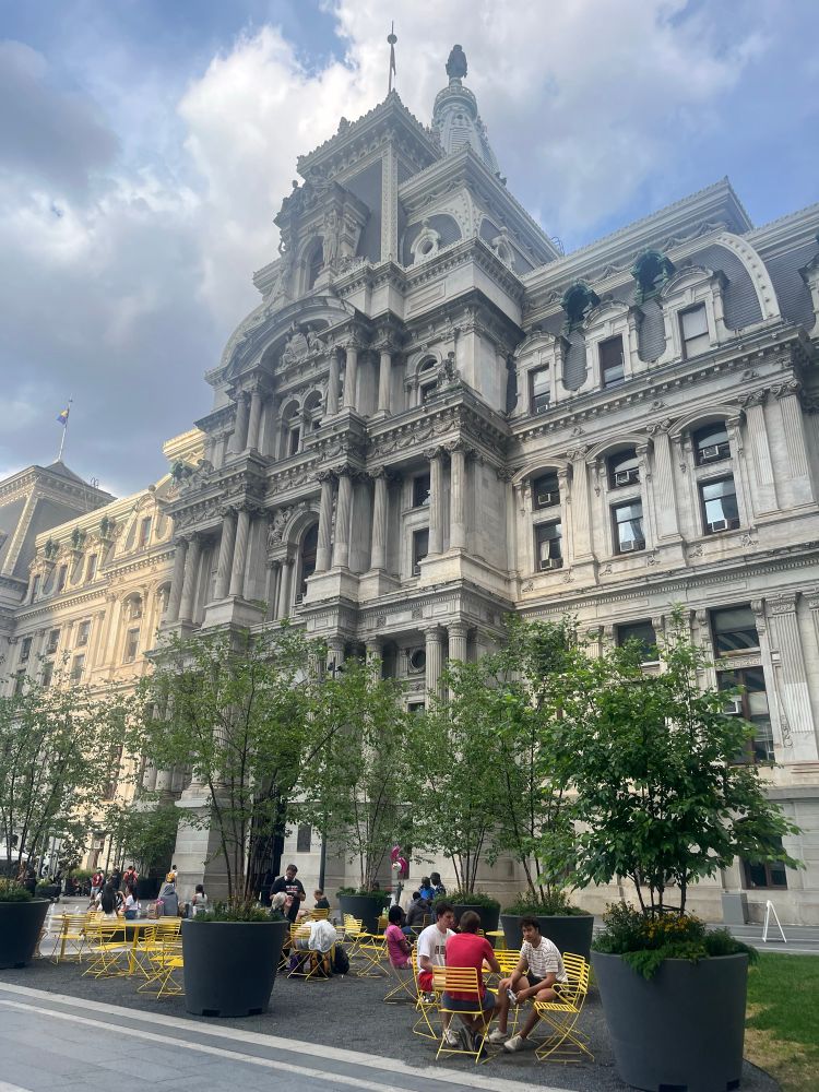 Photo of Philadelphia’s city hall building, 3 potted trees in front. Some small yellow tables with a few people sat