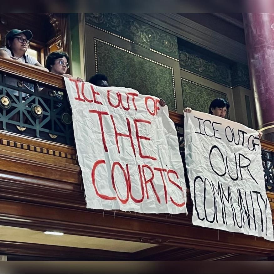 People stand on a balcony holding banners reading "ICE OUT OF THE COURTS" and "ICE OUT OF OUR COMMUNITY," expressing protest and solidarity.