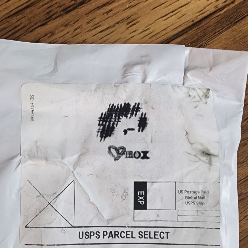Corner of a mailing package with the art by Mox icon, a typewriter-app made mini portrait of Mox's hair and one eye, underneath that a small hand drawn heart and it says mox. 