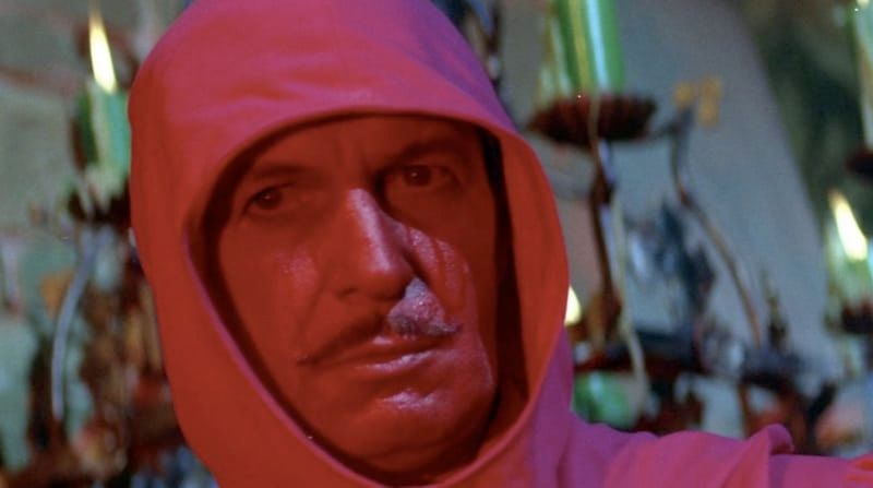 Man with a mustache with a blood red face wearing a red hood with candles and a stone wall in the background. 