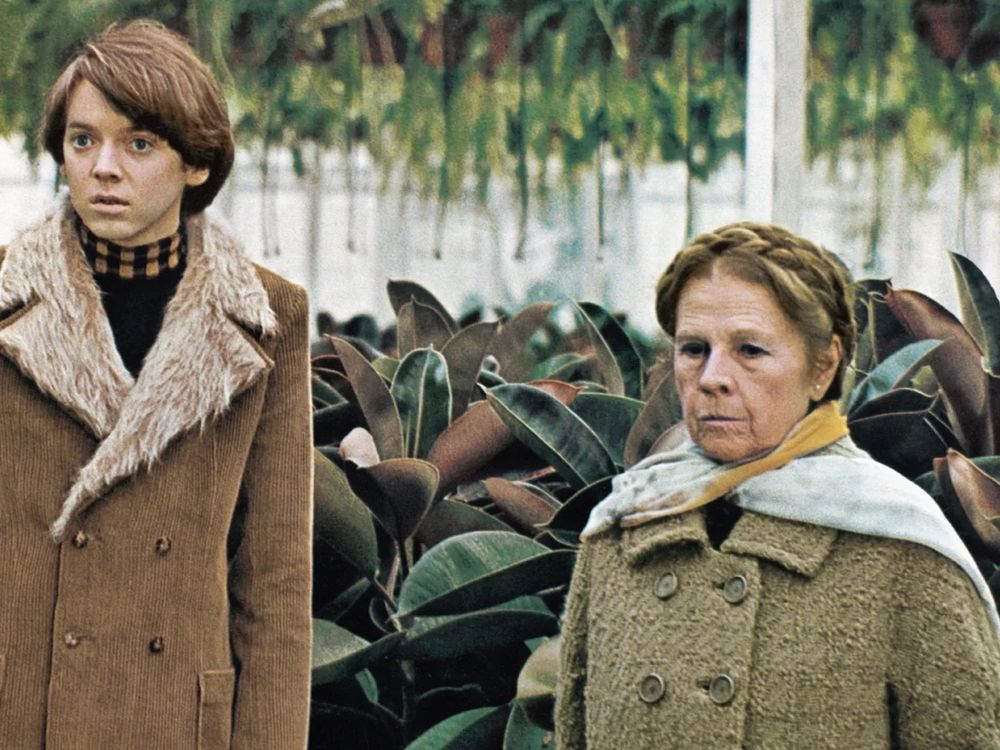 A young man with brown hair wearing a brown coat with furry lapels standing next to an elderly woman wearing a grey coat. They are standing in front of plants in a nursery in Redwood City, Calif. 