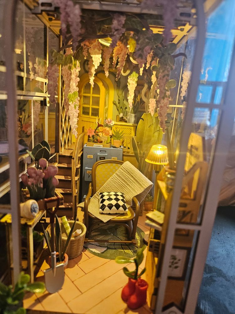 Close up of the house: You can see a rocking chair with a blanket and pillow, red boots, pictures, shelves, hanging flowers, and stairs up to a partially opened door.