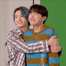 Jung Kook in a brown and white striped shirt with Grey hair has wrapped JHope in a hug. J Hope is in a brown and blue striped sweater and brown hair. Both are smiling.