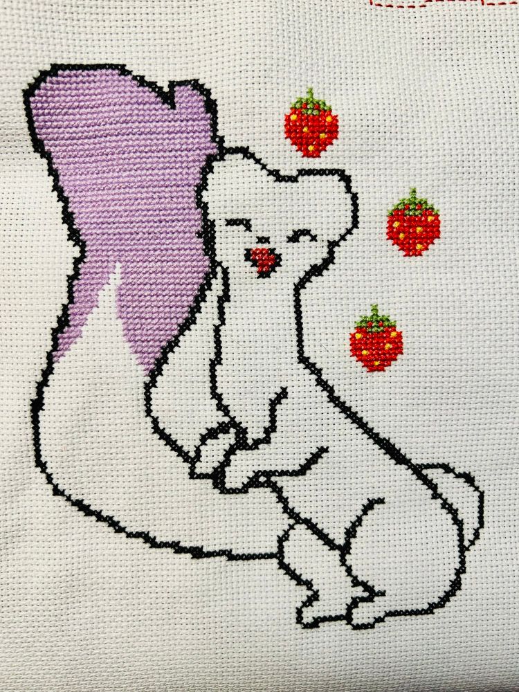 A cross stitch of Pip, a white stoat with a lavender-tipped tail. Two strawberries are floating on her right side and one is above her head