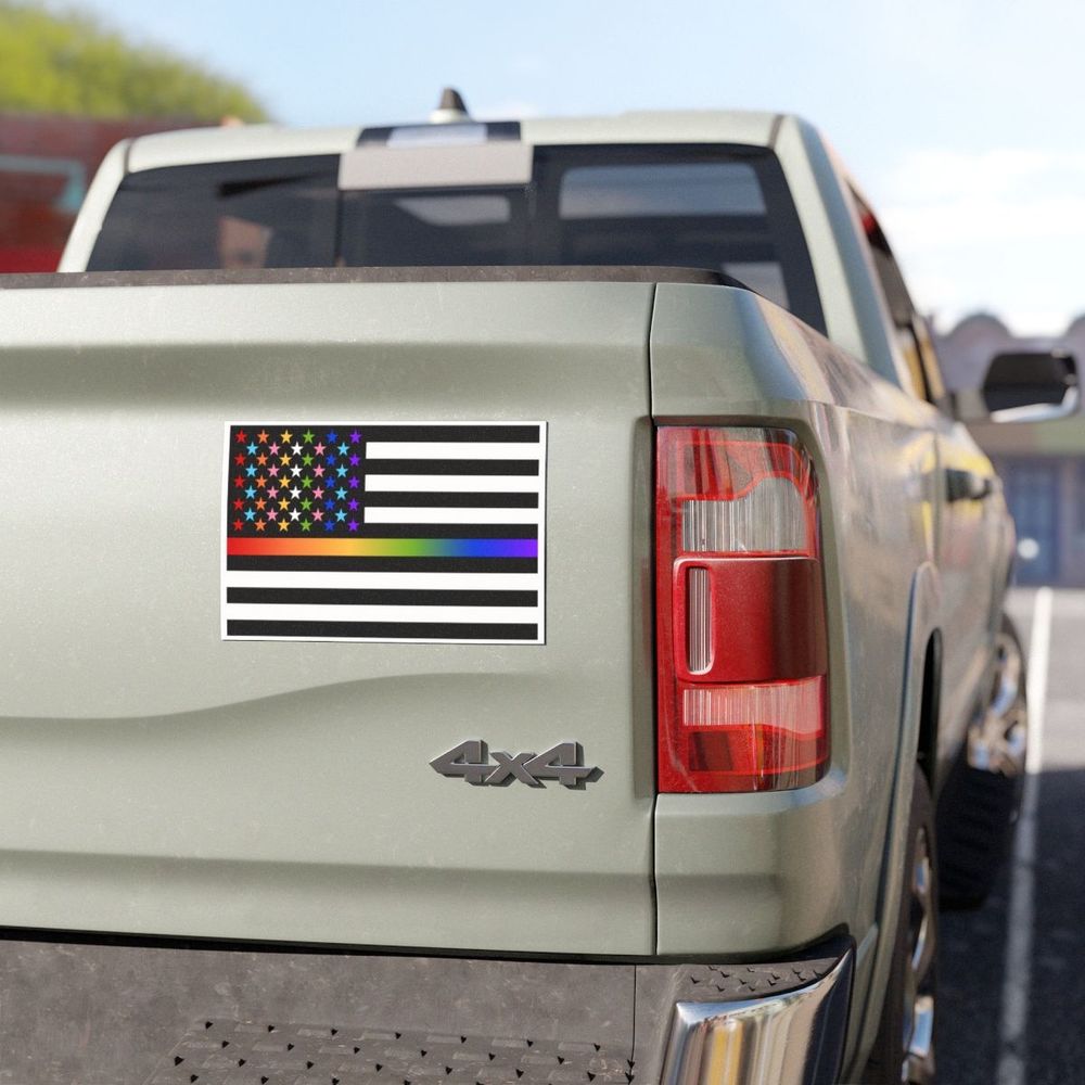 A large black, white, rainbow & trans pride colored 'Thin Queer Line' American flag magnet On the tailgate of a silver pickup truck.