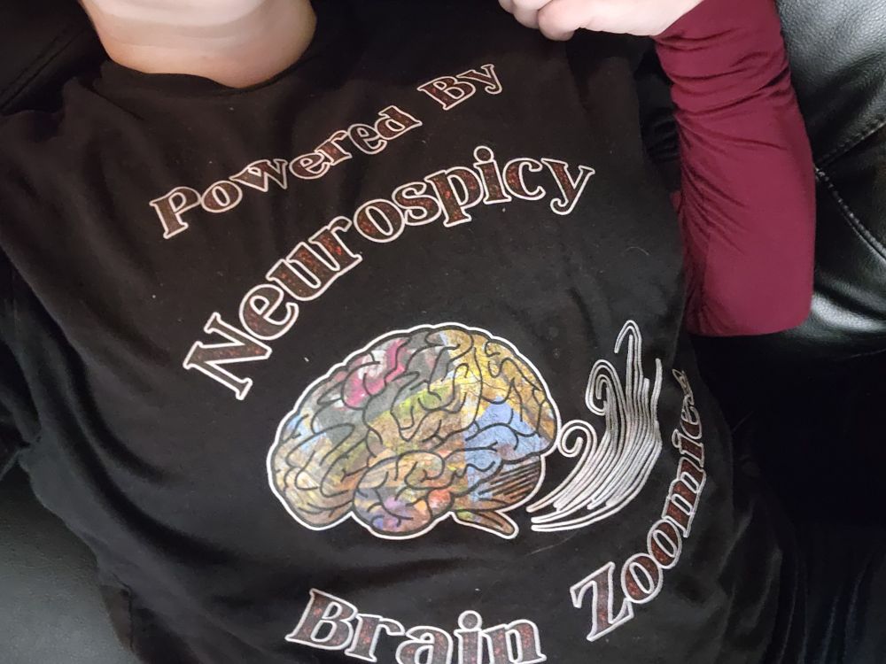 A tshirt that says "powered by neurospicy brain zoomies", with a large brain filled with photos that has little movement marks behind it. Inspired by a late night conversation about how it's when we try to finally sit down and relax that all the ideas hit.