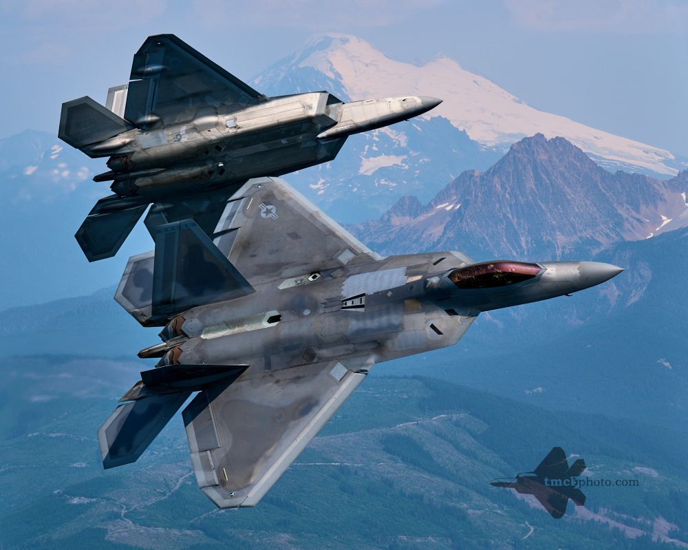 Two F-22 Raptors of the USAF F-22 Raptor Demo Team break formation with Mount Baker's glacier in the background. Both jets are facing the right of the image and mid turn, with the foreground jet revealing the topside of the F-22 as it turns towards the photo plane, and the background F-22 is partially obstructed by the foreground jet, with the bottomside visible as it pulls away from the photo plane.