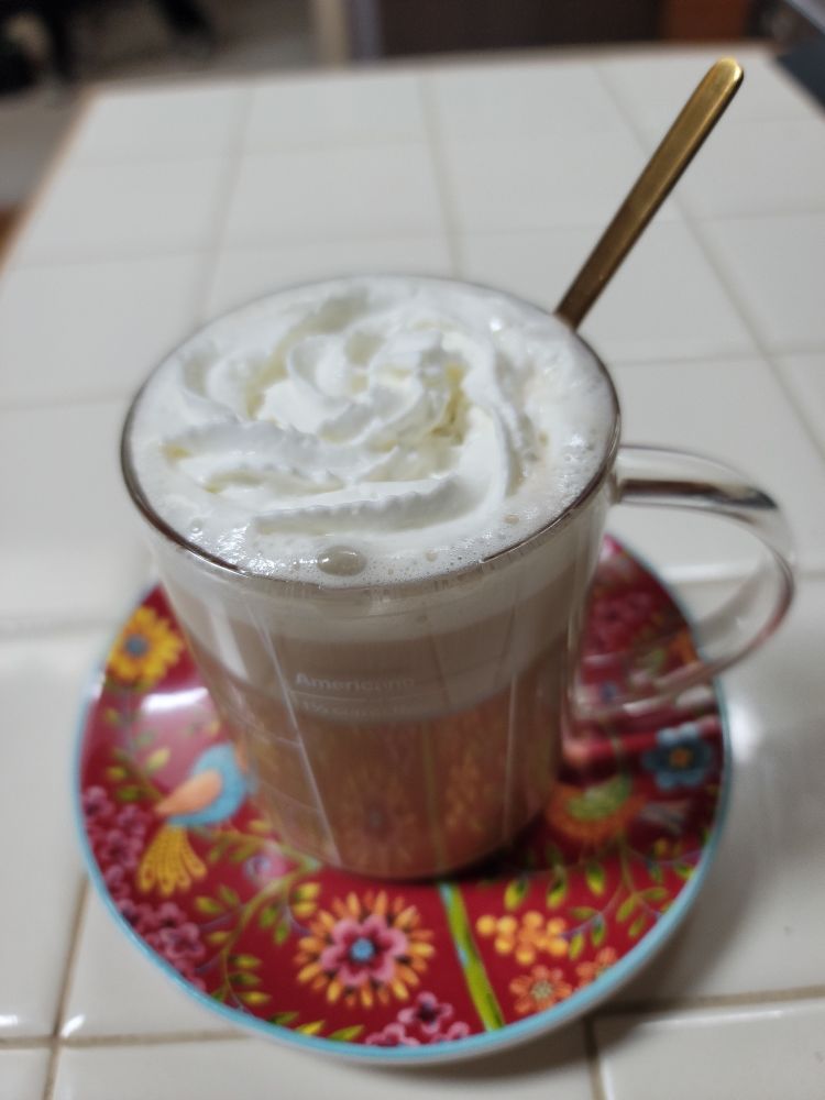 Photo of a clear glass mug of coffee, topped with whipped cream, sitting on a colorful red saucer with birds and flowers. 
