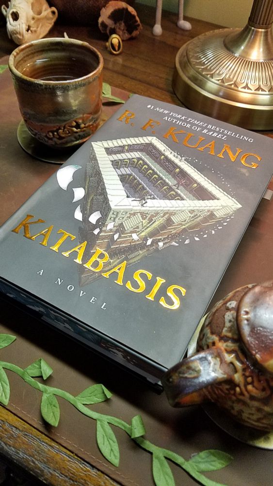 the book katabasis by r. f. kuang lays on a leather desk mat bordered by fabric leaves. next to it is a cup of steaming tea and a small tea pot, both mottled with warm glazes.