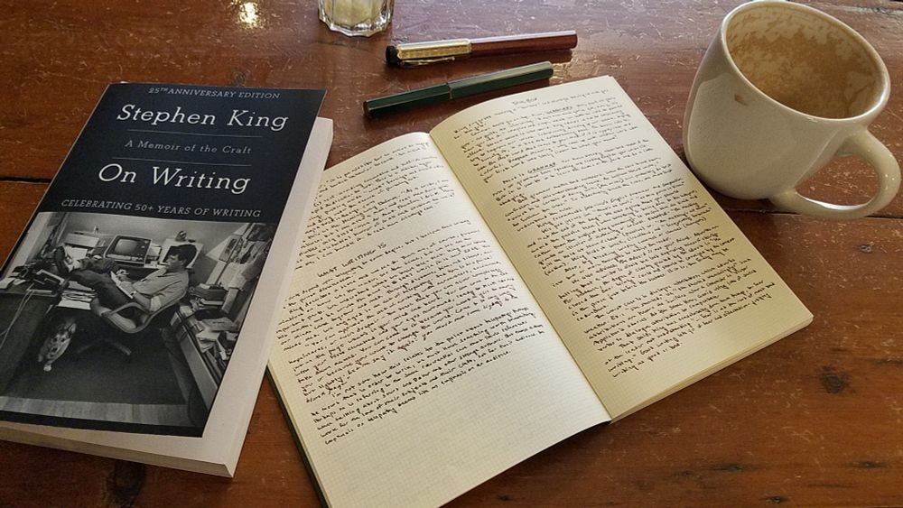 stephen king's on writing lays next to an open notebook full of tiny handwriting on a wooden table. two fountain pens and a near empty mug of latte rest nearby.