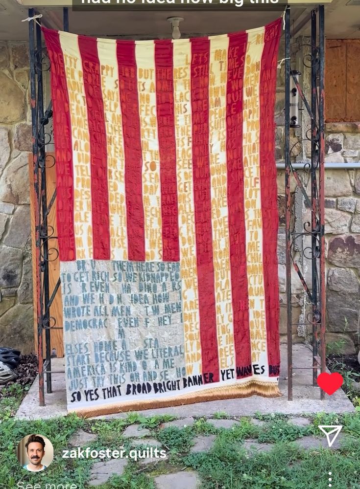 A quilt with the red & white stripes, & blue area, of the US flag. Quilted onto quilt are words telling how the US has always been a nation where some take from others. I believe in the link, which leads to ig, he voices what the quilt says. Zak is hanging this quilt on a house's outside entry way near front door. 