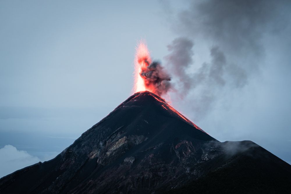 A photo of a volcano with a small explosion at its crater. The photo was taken in the early hours of the day, so the erupted material is incandescent with bright red and orange colors. Some of the red hot rocks released in the eruption appear to roll down the flank of the volcano. A small gray ash cloud accompanies the eruption.
