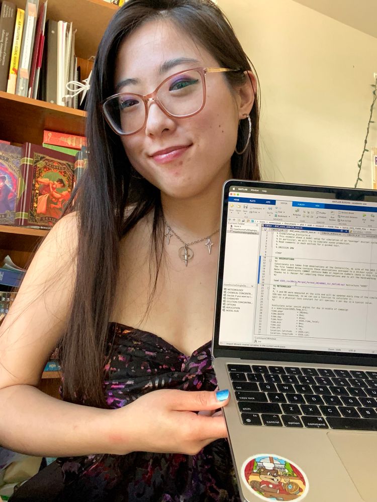 Author of tweet in the center of the photo, holding up her laptop. She wears a silver necklace with star and a sword charm, silver hoop earrings, with amber framed glasses, and a black and rainbow velvet strapless top